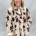 A smiling blonde woman wearing a cream-colored, long-sleeve button-up blouse with a black and red floral print, paired with black pants, set against a neutral gray studio background.
