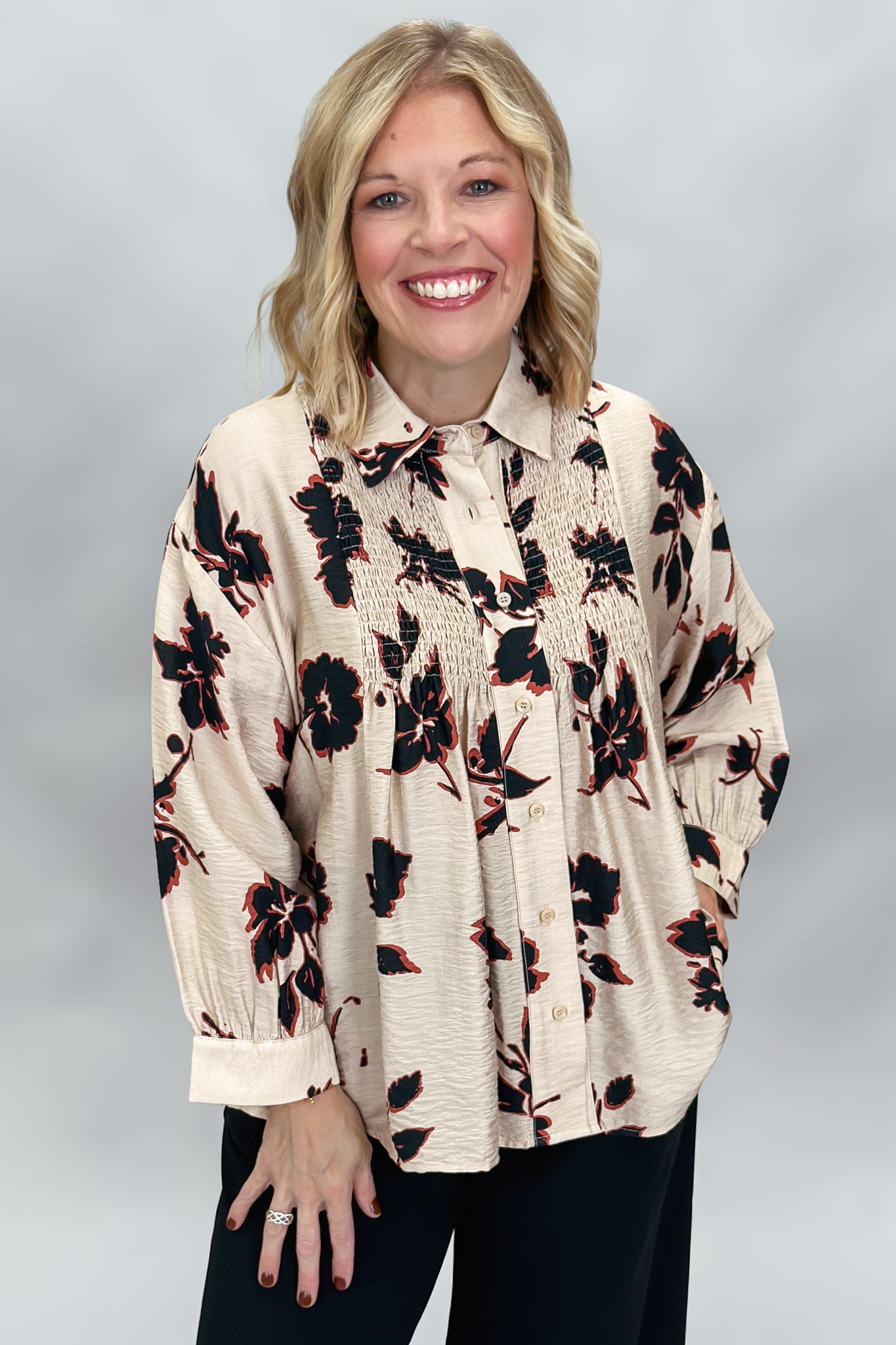 A smiling blonde woman wearing a cream-colored, long-sleeve button-up blouse with a black and red floral print, paired with black pants, set against a neutral gray studio background.