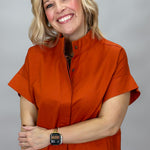 When Doves Cry orange mock-neck button-down shirt — A smiling blonde woman wearing a short-sleeve orange mock-neck button-up shirt and blue jeans, standing against a plain gray studio background.
