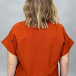 When Doves Cry orange mock-neck button-down shirt — A woman viewed from behind wearing a rust-orange short-sleeve top and blue jeans against a plain gray studio background.