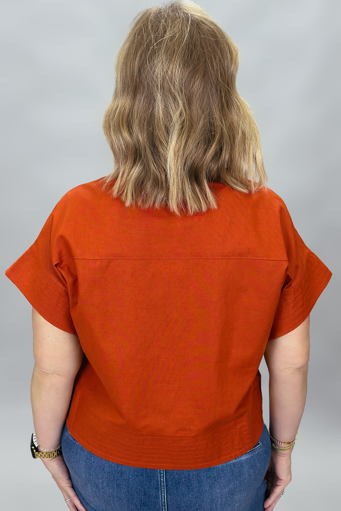 When Doves Cry orange mock-neck button-down shirt — A woman viewed from behind wearing a rust-orange short-sleeve top and blue jeans against a plain gray studio background.