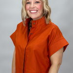 When Doves Cry orange mock-neck button-down shirt — A smiling blonde woman wearing a bright orange short-sleeve zip-front jacket with a stand collar, paired with blue jeans, photographed against a neutral gray studio background.