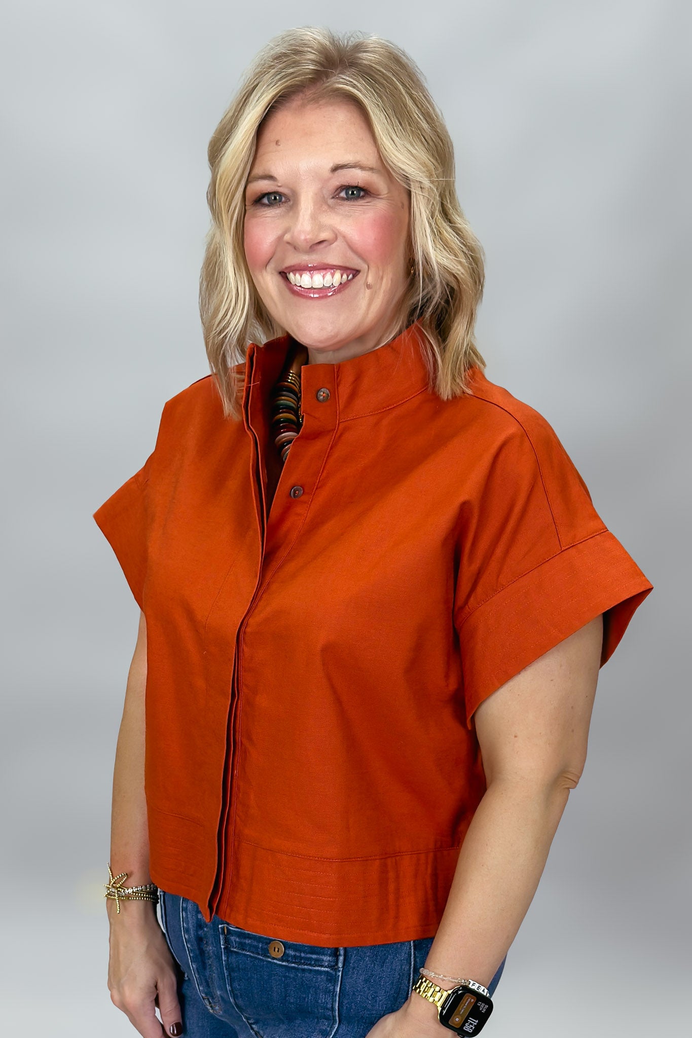 When Doves Cry orange mock-neck button-down shirt — A smiling blonde woman wearing a bright orange short-sleeve zip-front jacket with a stand collar, paired with blue jeans, photographed against a neutral gray studio background.