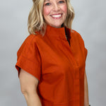 When Doves Cry orange mock-neck button-down shirt — A smiling blonde woman wearing a short-sleeve orange mock-neck top and blue jeans, standing against a light gray studio background.