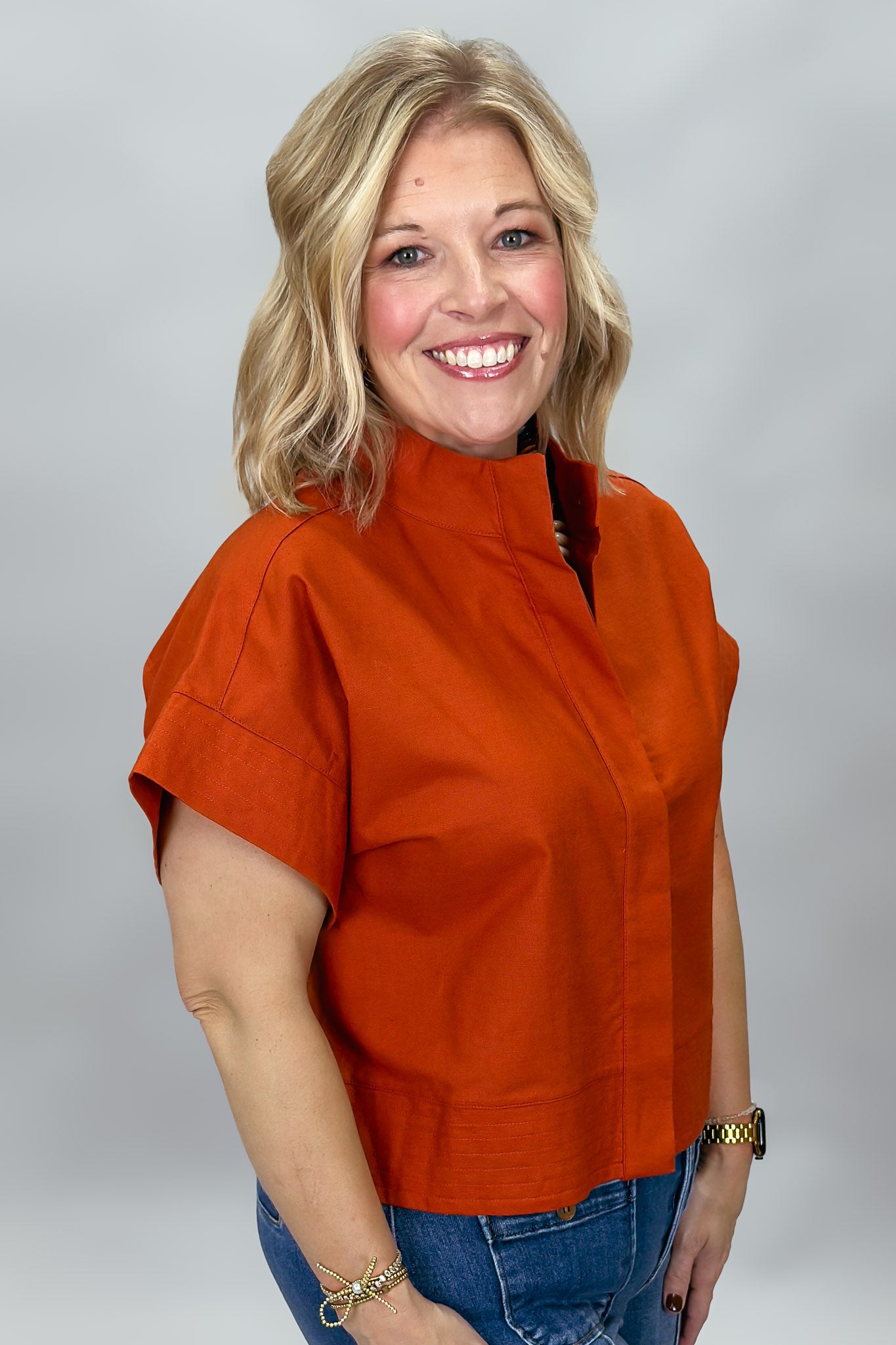 When Doves Cry orange mock-neck button-down shirt — A smiling blonde woman wearing a short-sleeve orange mock-neck top and blue jeans, standing against a light gray studio background.
