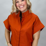 When Doves Cry orange mock-neck button-down shirt — A smiling blonde woman wearing a short-sleeve orange button-front shirt with a stand collar and blue jeans, photographed against a plain light gray studio background.