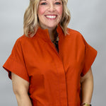 When Doves Cry orange mock-neck button-down shirt — A smiling blonde woman wearing a bright orange short-sleeve button-front top with a stand collar and blue jeans, photographed against a light gray studio background.