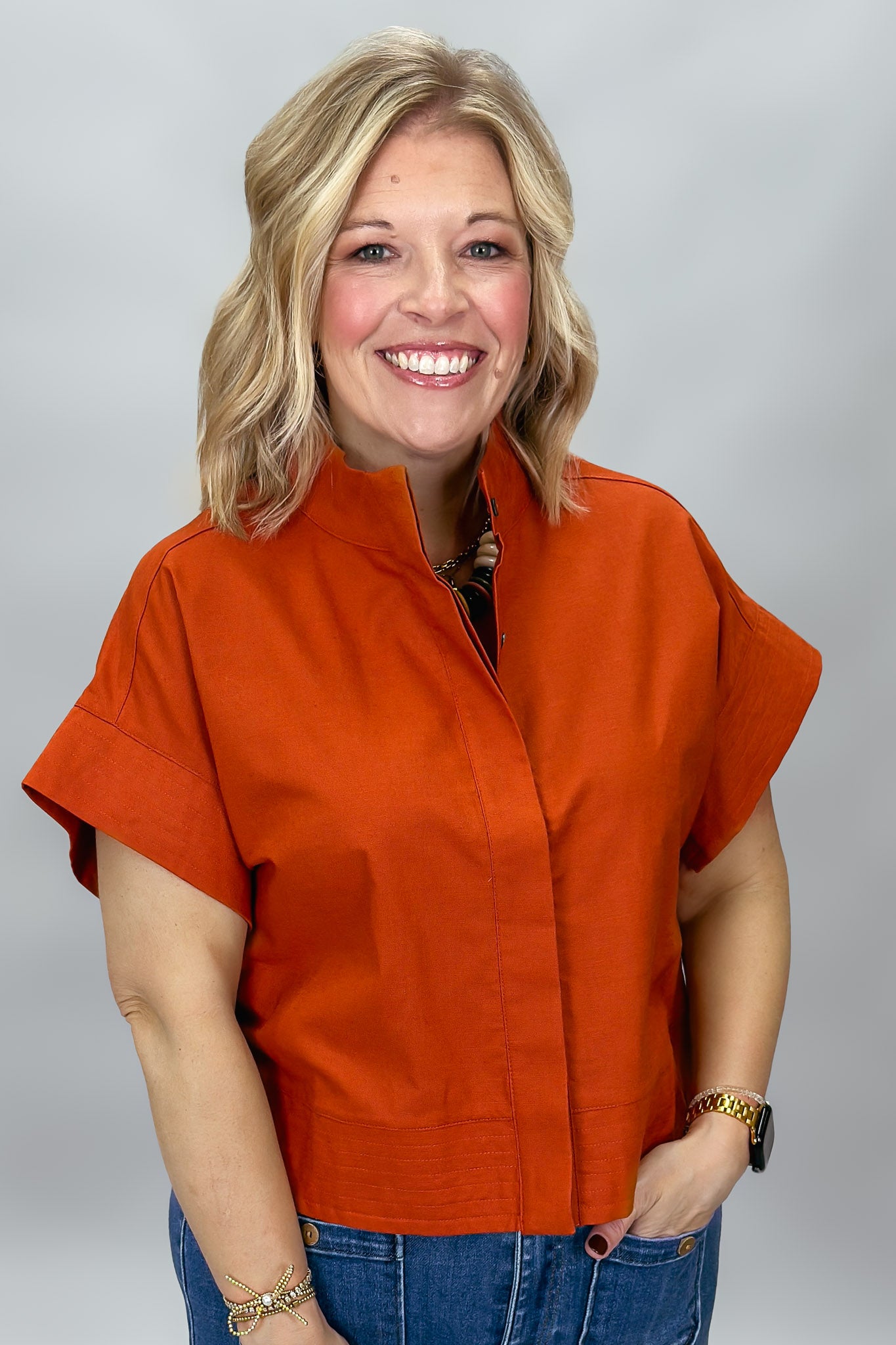 When Doves Cry orange mock-neck button-down shirt — A smiling blonde woman wearing a bright orange short-sleeve button-front top with a stand collar and blue jeans, photographed against a light gray studio background.